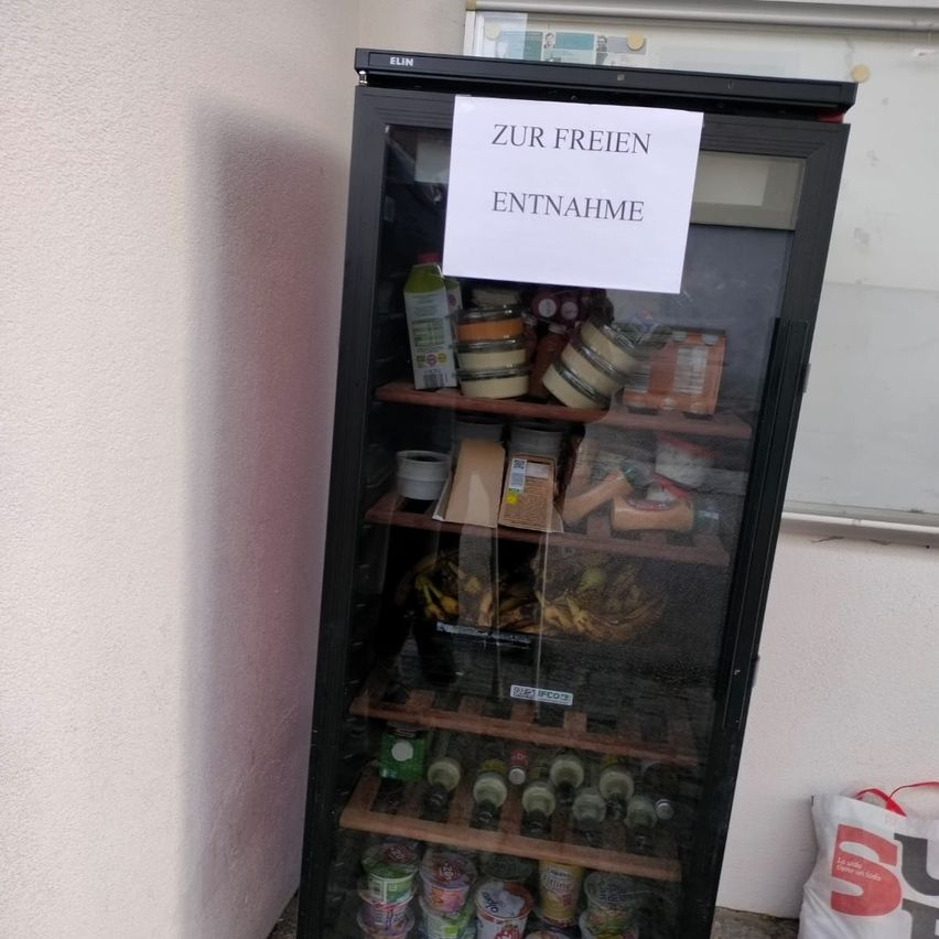 Ein Kühlschrank mit einem Schild oben, das 'ZUR FREIEN ENTNAHME' besagt. Der Kühlschrank ist mit verschiedenen Lebensmitteln bestückt.
