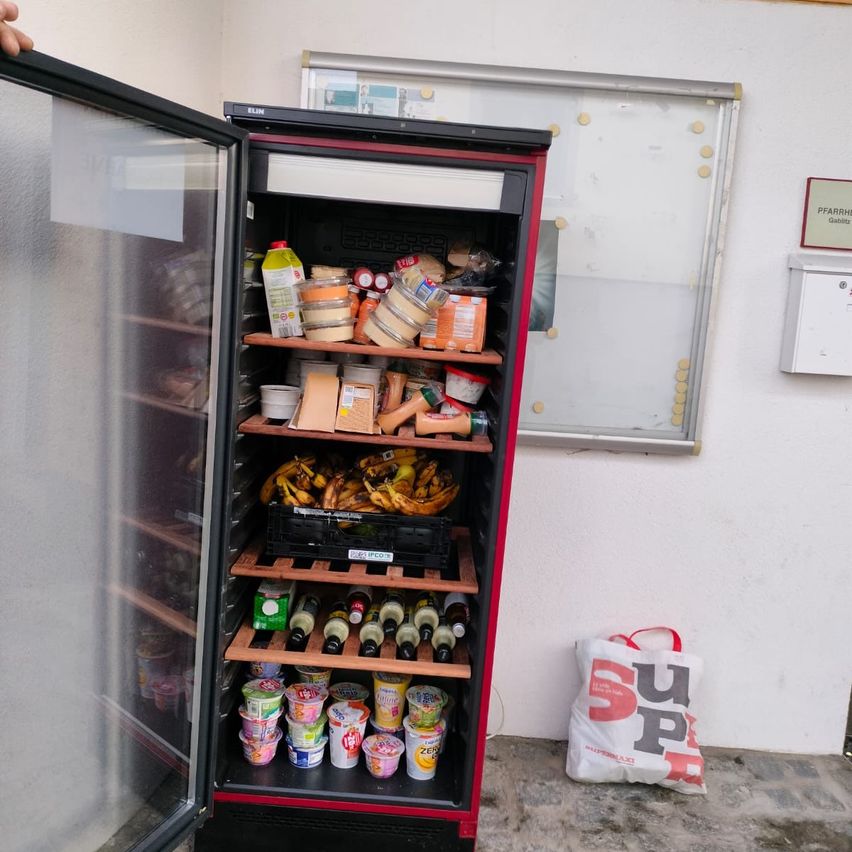 Ein offener Kühlschrank mit Regalen voller Lebensmittel, darunter Bananen, Joghurt und Flaschen. Daneben steht ein weißer Beutel mit der Aufschrift 'SUP'.