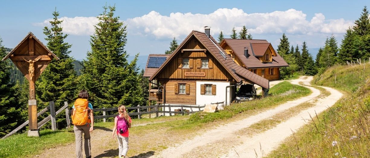 Zwei Wanderer, einer mit Rucksack und der andere mit kleinem Rucksack, gehen auf einem Feldweg mit einem Haus im Hintergrund.