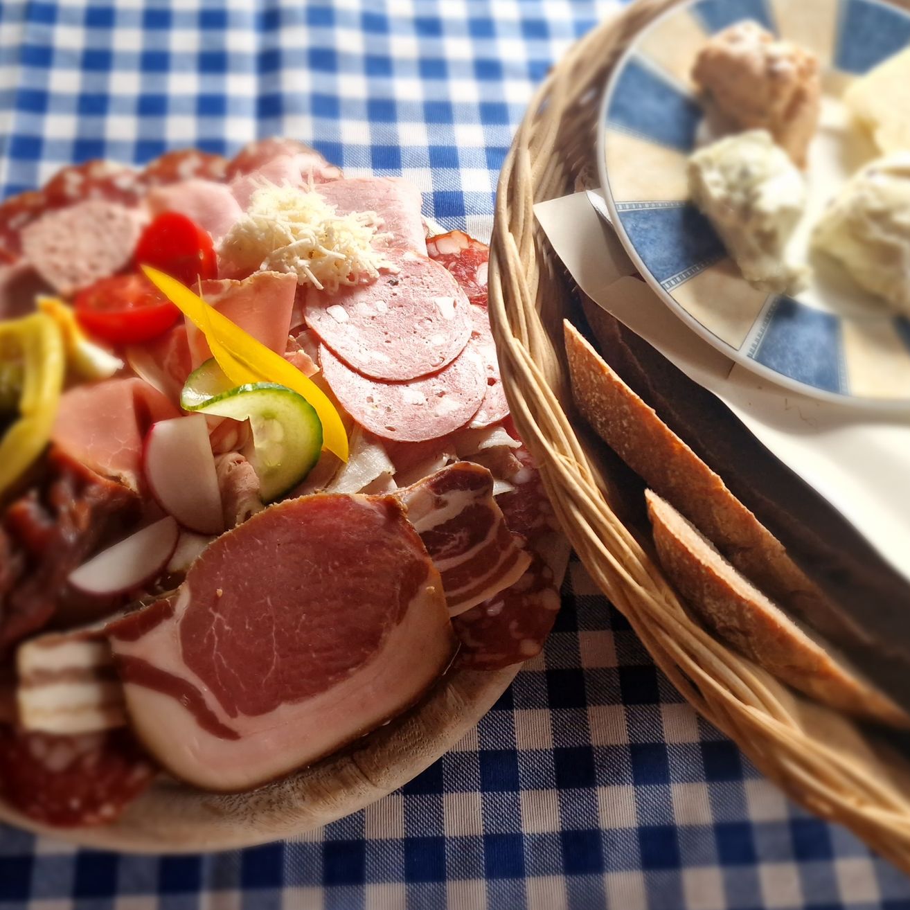 Ein Tablett mit Fleisch, Käse und Gemüse auf einem blau-weiß karierten Tischtuch, begleitet von einem Korb mit Brot und einem Teller mit Beilagen.