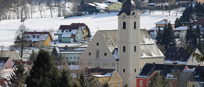 Ein verschneites Dorf im Winter mit einem hohen weißen Kirchturm. Die Häuser sind in verschiedenen Farben gestrichen, einige mit Schnee auf den Dächern. Die Landschaft ist umgeben von Bäumen und Bergen.