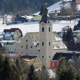 Eine Winterszene eines Dorfes mit schneebedeckten Dächern, einem hohen Kirchturm und einem Uhrturm. Verschiedene Häuser in verschiedenen Farben säumen die verschneite Landschaft.