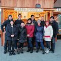 Eine Gruppe von Menschen posiert für ein Foto vor einem hölzernen Kiosk mit der Aufschrift 'Gruppe Prosecco'. Sie tragen Winterkleidung und einige haben Mützen auf.
