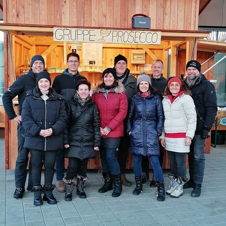 Eine Gruppe von Menschen posiert für ein Foto vor einem hölzernen Kiosk mit der Aufschrift 'Gruppe Prosecco'. Sie tragen Winterkleidung und einige haben Mützen auf.
