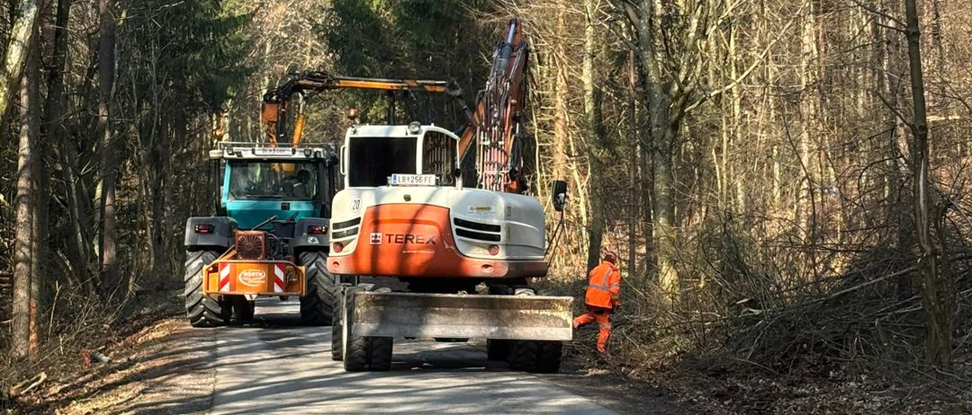 Zwei schwere Baufahrzeuge stehen auf einer Straße im Wald. Eines ist orange und weiß, das andere blau. Ein Arbeiter in einer orangefarbenen Sicherheitsjacke steht in der Nähe.