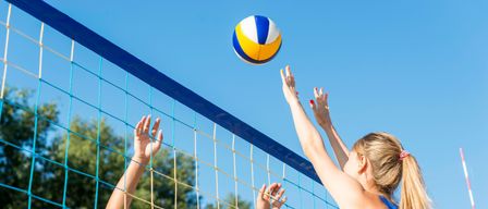 Zwei Frauen spielen Volleyball, eine greift nach dem Ball, der über das Netz fliegt. Die Szene spielt sich im Freien ab.