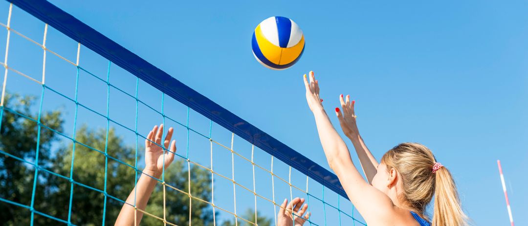 Zwei Frauen spielen Volleyball, eine greift nach dem Ball, der über das Netz fliegt. Die Szene spielt sich im Freien ab.