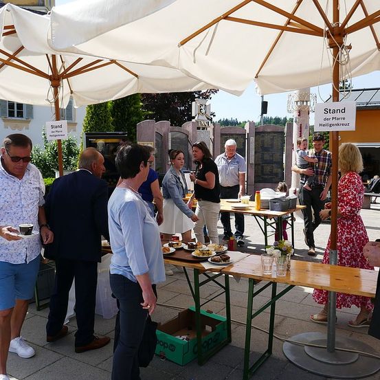 Mehrere Menschen stehen unter Schirmen, möglicherweise bei einem Treffen. Ein Tisch mit Essen ist aufgebaut, und Menschen stehen darum herum. Eine Frau in einem Blumenkleid steht rechts. Ein Mann hält ein Baby und steht neben einem anderen Mann. Dahinter steht ein Gedenkstein.