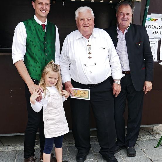 Drei Männer und ein junges Mädchen stehen zusammen draußen. Die Männer tragen formelle Kleidung, und das Mädchen hält ein Papierschild. Der Mann in der Mitte trägt eine Uhr und einen Gürtel. Hinter ihnen ist eine Wand mit etwas Text.