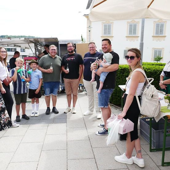 Eine Gruppe von Erwachsenen und Kindern posiert für ein Foto vor einem Gebäude. Eine Frau hält ein Baby und eine Tasche. Zwei Männer in der Mitte halten Flaschen. Eine Frau auf der rechten Seite trägt eine Sonnenbrille.