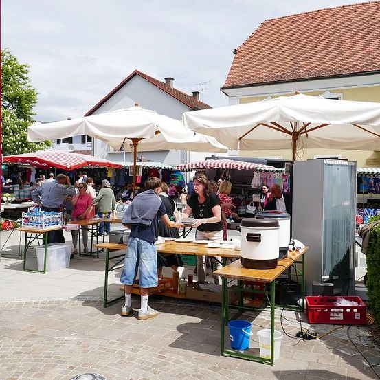 Ein Markt im Freien mit mehreren Ständen unter weißen Sonnenschirmen. Ein Stand hat einen Mann, der einer Frau Essen serviert. Töpfe stehen auf einem Tisch und Eimer in der Nähe. Menschen kaufen ein und Gebäude mit roten Dächern sind im Hintergrund.