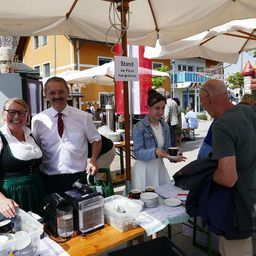 Vier Personen, zwei Männer und zwei Frauen, stehen an einem Tisch unter einem Schirm mit einem Schild, auf dem 'Stand der Pfarre Heiligenkreuz' steht. Die Frau in der Mitte hält eine Tasse. Der Tisch hat Teller, Tassen und eine Kaffeemaschine. Im Hintergrund sind Menschen, Schirme und Gebäude zu sehen.