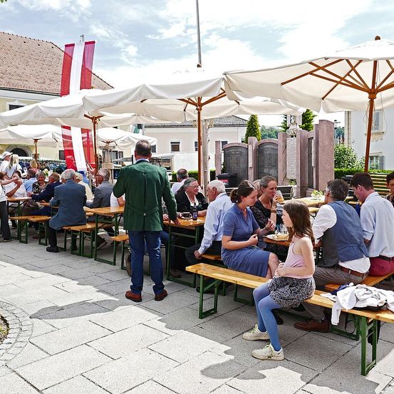Eine Gruppe von Menschen sitzt unter Schirmen in einem Essbereich im Freien. Einige haben Gläser, Teller und Flaschen auf den Tischen. Ein Mann steht in der Nähe der Tische. In der Ferne befinden sich Gebäude und eine Flagge.