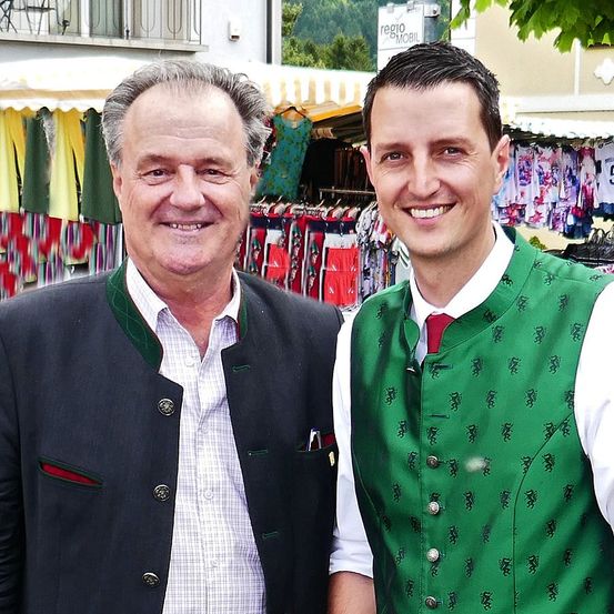 Zwei Männer stehen nebeneinander und lächeln, möglicherweise auf einem Marktplatz. Sie tragen traditionelle Kleidung. Der Mann auf der linken Seite hat graues Haar und trägt einen schwarzen Mantel. Der Mann auf der rechten Seite trägt eine grüne Weste und eine rote Krawatte.