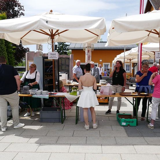 Menschen versammeln sich um einen Essensstand mit Schirmen. Eine Frau in einem weißen Kleid steht vor dem Tisch, während andere auf ihren Handys sprechen. Im Hintergrund befinden sich Pflanzen und Bäume.