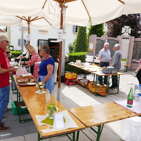 Eine Gruppe von Menschen bereitet sich auf eine Veranstaltung im Freien vor. Zwei lange Tische sind unter Sonnenschirmen aufgestellt, mit Essen und Getränken darauf. Ein Gebäude mit einer Statue im Hintergrund ist sichtbar.