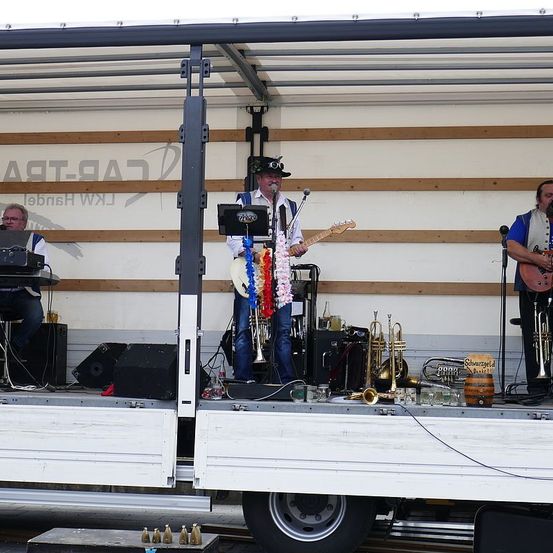 Eine Band spielt auf einer Bühne, die aus einem Lastwagen besteht, mit einem Gitarristen, einem Keyboarder und einem Trompeter. Dahinter ist eine Holzwand mit einem Logo zu sehen.