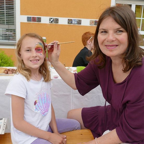 Eine Frau malt einer jungen Mädchen auf der Bank Gesichtsfarbe auf. Das Mädchen trägt ein weißes T-Shirt mit einer bestickten Muschel. Sie sind in einer Außenumgebung mit einem Tisch und einer Person im Hintergrund.