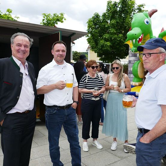 Vier Personen stehen im Freien, möglicherweise auf einem Festival. Sie halten Getränke in den Händen. Hinter ihnen steht ein Gebäude, ein Baum und ein großer aufblasbarer Drache.