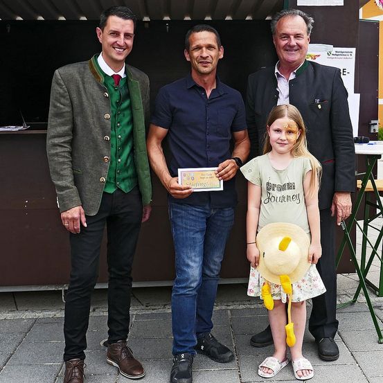 Drei Männer und ein junges Mädchen stehen vor einem Stand. Der Mann in der Mitte hält ein Zertifikat. Der Mann auf der linken Seite trägt eine grüne Weste. Der Mann auf der rechten Seite trägt einen schwarzen Anzug. Das Mädchen trägt einen Blumenrock und einen Hut.