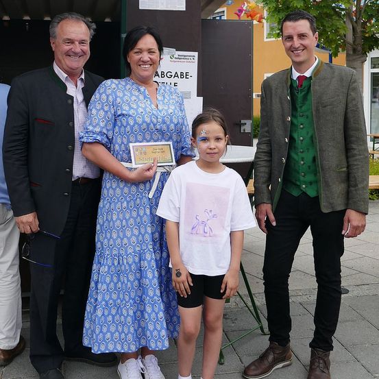 Vier Personen posieren für ein Foto, zwei Erwachsene, einer in einem traditionellen Anzug und der andere in einem blauen Kleid, sowie zwei Kinder, eines mit Gesichtsbemalung. Sie stehen draußen mit einem Schild, das 'Abgabe' steht.