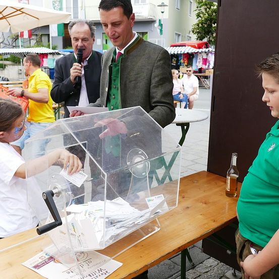 Ein Mann spricht in ein Mikrofon, während er neben einer Wahlurne steht, eine junges Mädchen greift hinein. Ein weiterer Mann steht hinter der Urne, und Menschen sitzen auf einer Bank in der Nähe. Eine Flasche steht auf dem Tisch.