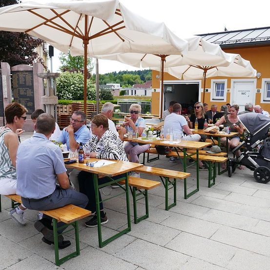 Eine Gruppe von Menschen sitzt an Holztischen in einem Café im Freien und genießt ihre Mahlzeiten und Getränke unter großen Sonnenschirmen.