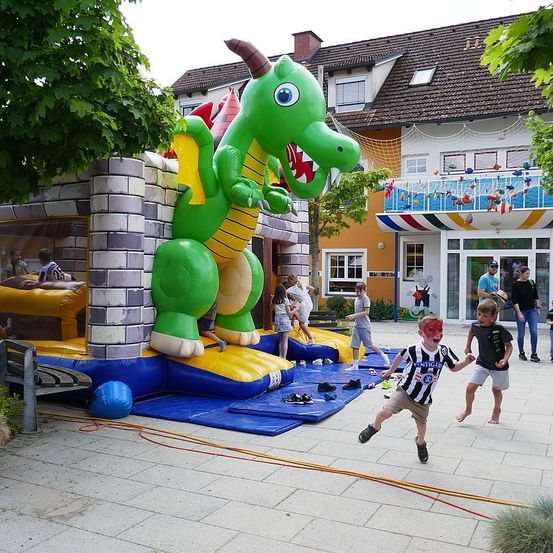 Kinder spielen in einem Innenhof um eine aufblasbare Drachenburg. Einige springen hinein und hinaus aus der Burg.