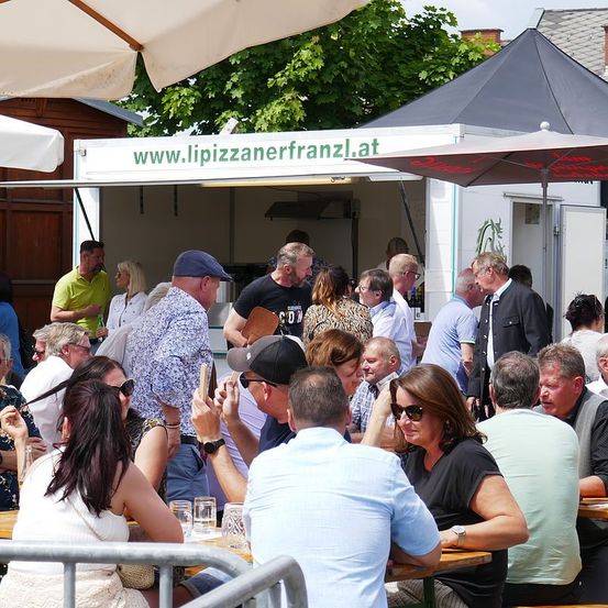 Menschen versammeln sich um Tische unter Sonnenschirmen in einem Outdoor-Restaurant, möglicherweise einem Food-Truck. Einige essen, während andere in Gespräche vertieft sind. Der Food-Truck zeigt eine Website-Adresse an.