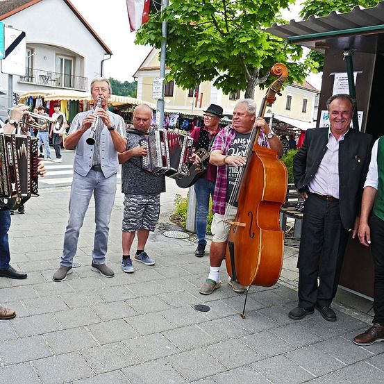 Eine Gruppe von Musikern spielt Instrumente im Freien. Sie bestehen aus einem Mann, der Klarinette spielt, einem anderen mit einem Akkordeon, einem Cellisten und einem Trompeter. Sie stehen vor einem Baum mit einer Flagge.