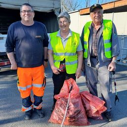 Drei Personen in Warnwesten stehen neben einem Lastwagen und Säcken mit Müll. Der Mann auf der linken Seite trägt eine orangefarbene Arbeitskleidung. Die Frau in der Mitte lächelt und der Mann auf der rechten Seite hält einen Stock in der Hand und schaut nach vorne.