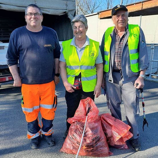 Drei Personen in Warnwesten stehen neben einem Lastwagen und Säcken mit Müll. Der Mann auf der linken Seite trägt eine orangefarbene Arbeitskleidung. Die Frau in der Mitte lächelt und der Mann auf der rechten Seite hält einen Stock in der Hand und schaut nach vorne.