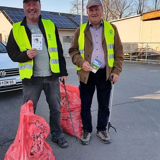 Zwei Männer stehen in einem Parkplatz, lächeln und tragen Warnwesten. Einer hält einen Müllsack und einen Stock, der andere ein Büchlein. Ein Auto mit dem Kennzeichen '300M 98' ist hinter ihnen geparkt.