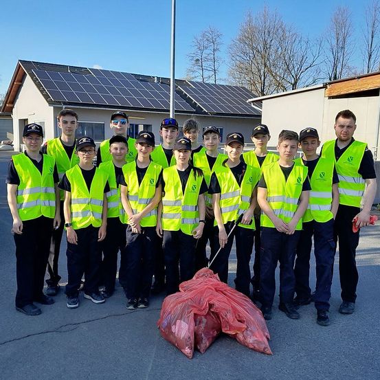 Eine Gruppe junger Freiwilliger in neongrünen Warnwesten posiert vor einem Gebäude mit Solarmodulen und hält einen roten Müllsack.