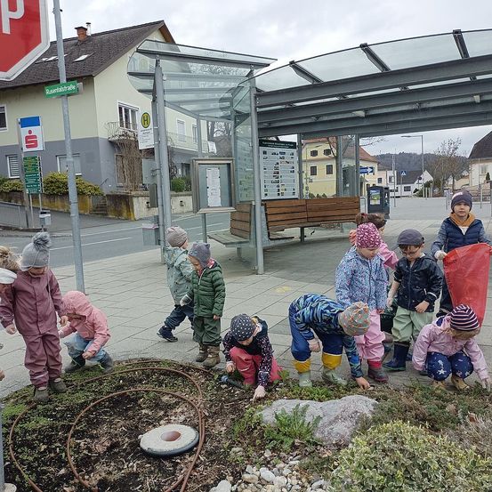 Kinder in Winterkleidung versammeln sich um ein Blumenbeet, möglicherweise an einer Bushaltestelle, mit einem Stoppschild und einem Haus im Hintergrund.