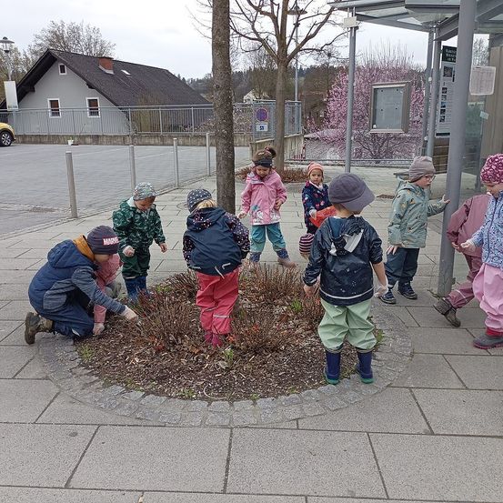 Eine Gruppe von Kindern ist draußen und einige pflanzen um einen Baum herum. Sie tragen warme Kleidung und Stiefel. Dahinter steht ein Haus mit einem Zaun und einem Baum mit rosa Blüten.