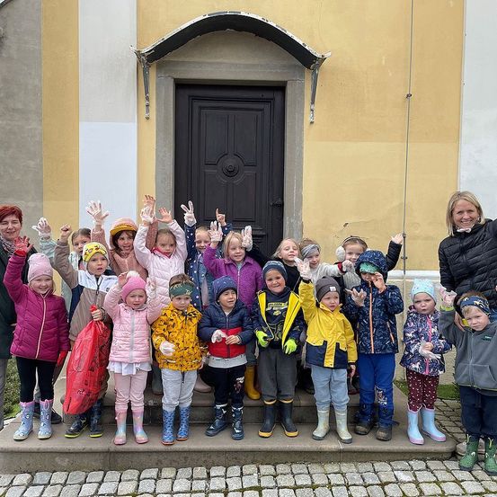 Eine Gruppe von Kindern, Erwachsenen und einer Frau posiert für ein Foto vor einem gelben Gebäude mit einer schwarzen Tür. Sie tragen alle Winterkleidung und einige haben Stiefel an. Die Frau lächelt.