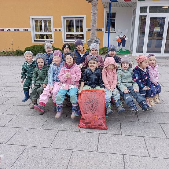 Eine Gruppe von Kindern, alle in Winterkleidung gekleidet, sitzen zusammen auf einer Bank vor einem Gebäude mit gelben Wänden und Glastüren.