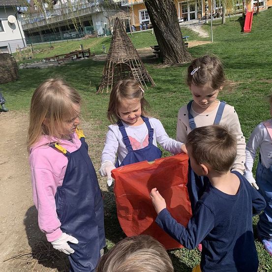 Vier Kinder spielen draußen. Zwei Mädchen und ein Junge halten ein rotes Netz, während das andere Mädchen zusieht. Dahinter steht ein Haus und ein Spielplatz.