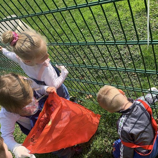 Kinder sind draußen an einem Zaun, eines hält eine rote Tasche, ein anderes schaut hinein, und ein Junge mit Rucksack in der Nähe.