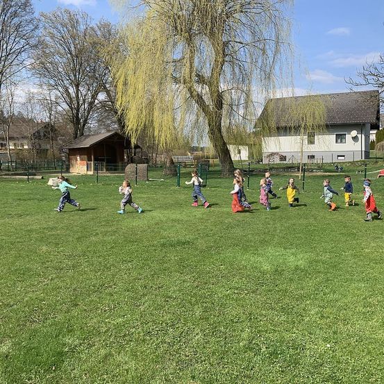 Kinder spielen auf einer Grasfläche, einige in Stiefeln, mit Häusern und einem Baum im Hintergrund.