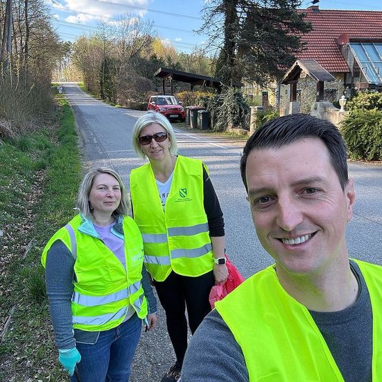 Drei Personen in Sicherheitswesten und Handschuhen stehen auf einer Straße. Dahinter befindet sich ein Haus mit rotem Dach, ein rotes Auto und ein Mülleimer.