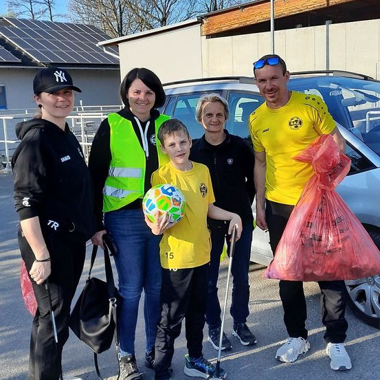 Fünf Personen, darunter ein Junge mit einem Fußball, posieren für ein Foto neben einem Auto. Zwei Frauen tragen Taschen, und eine weitere hält einen Stock. Dahinter befindet sich ein Gebäude mit Solarmodulen.