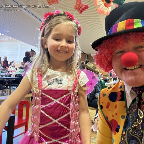 Ein lächelndes junges Mädchen in einem rosa Kleid mit Blumenkranz posiert neben einem Clown in einem bunten Hut mit roter Nase. Sie sind drinnen, möglicherweise bei einer Kinderveranstaltung.