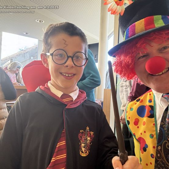 Ein Junge, verkleidet als Harry Potter mit einem Zauberstab, lächelt, neben einem Clown mit roter Nase.