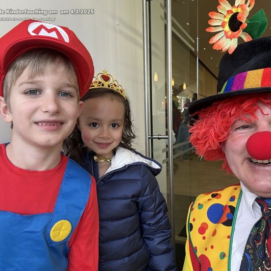 Ein Junge und ein Mädchen in Kostümen posieren mit einem Clown in einem farbenfrohen Kostüm, lächeln in die Kamera. Der Clown hat eine rote Nase und eine Blume auf seinem Hut.