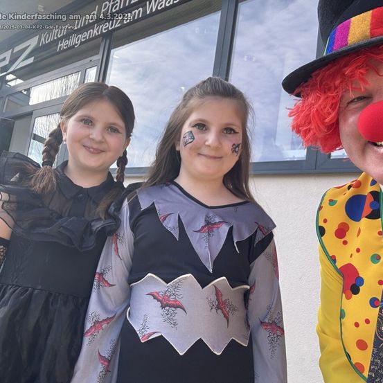 Drei Kinder in Halloween-Kostümen stehen vor einem Gebäude. Ein Clown in bunter Kleidung steht neben ihnen. Das Mädchen in der Mitte hat Gesichtsbemalung auf den Wangen.