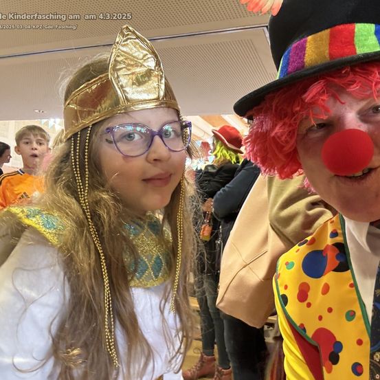 Ein Foto vom Kinderfasching am 4. März 2025. Ein Mädchen in einer goldenen Kopfbedeckung und Brille lächelt in die Kamera, neben einem Clown mit roter Nase. Hinter ihnen sind Menschen in verschiedenen Kostümen zu sehen, darunter eine Person mit rotem Hut.