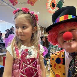 Ein junges Mädchen in einem Feenkostüm posiert neben einem Clown mit roter Nase bei einem Kinderfasching. Der Hintergrund zeigt einen Raum mit Menschen und Dekorationen.