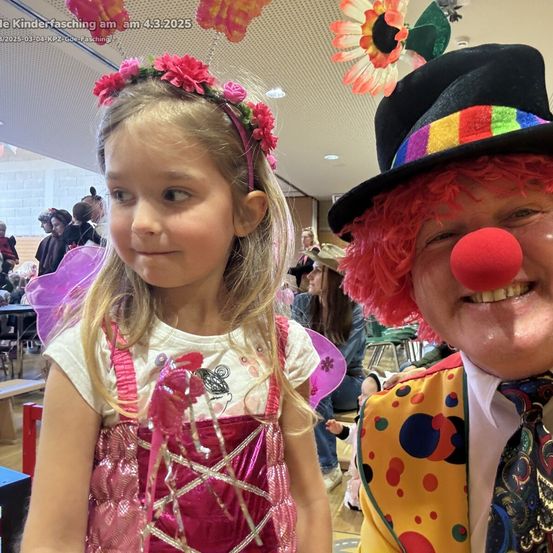 Ein junges Mädchen in einem Feenkostüm posiert neben einem Clown mit roter Nase bei einem Kinderfasching. Der Hintergrund zeigt einen Raum mit Menschen und Dekorationen.